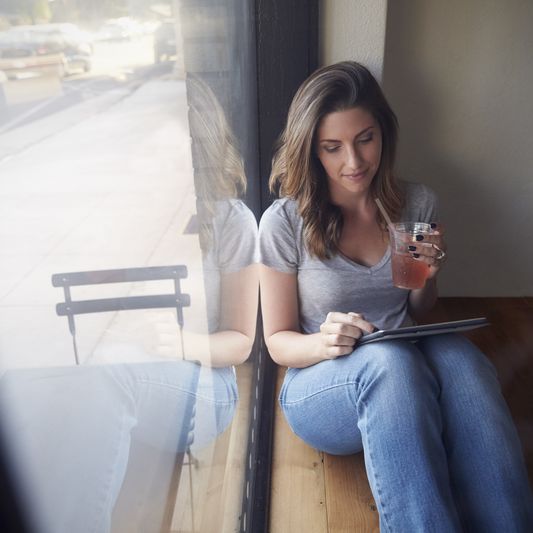 A young woman checks her iPad while sitting in a window.